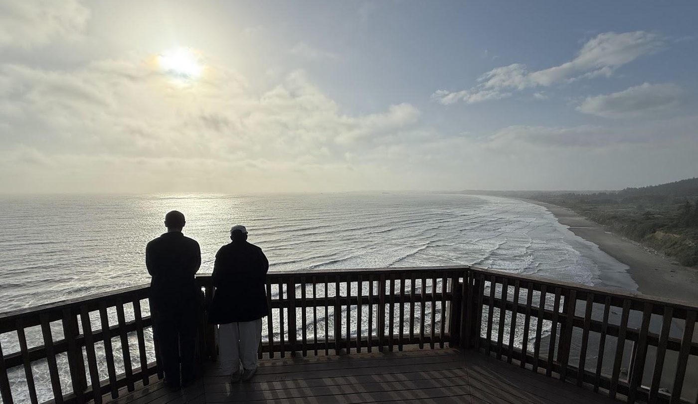 Two figures at a wooden overlook, watching the sun over the Pacific and a long curve of coastline.