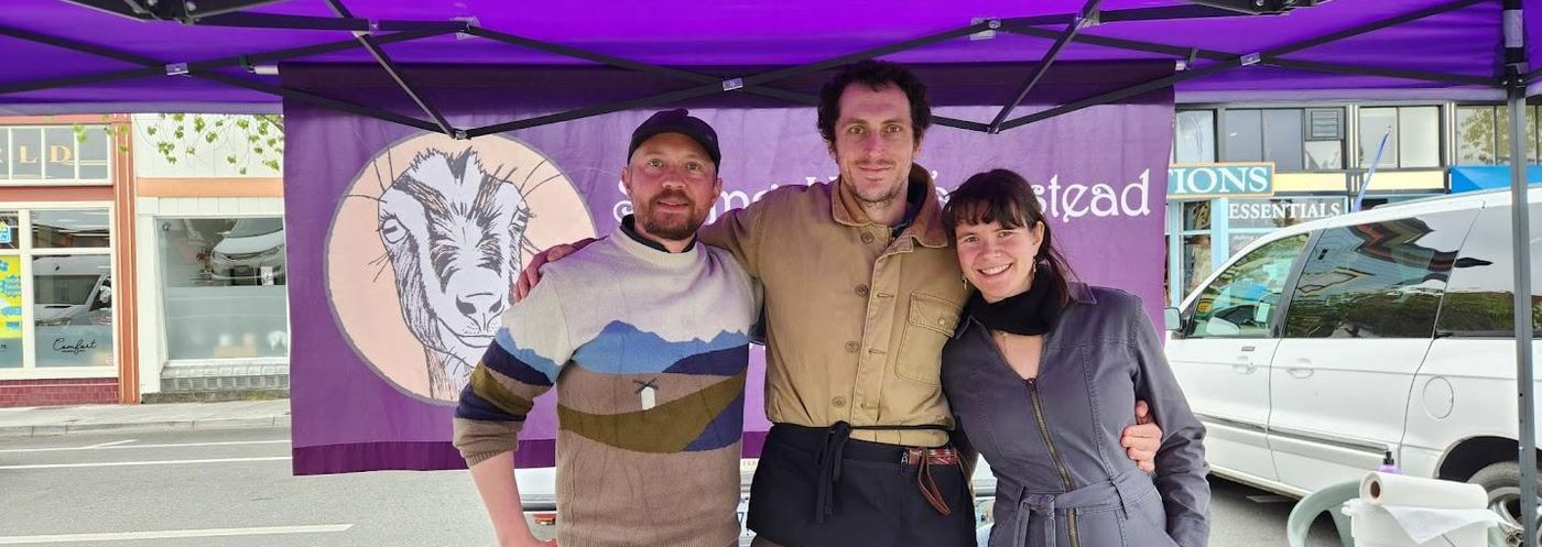 Three visitors smiling at a goat's cheese stall under a purple canopy at a street market.